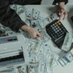 Jak liczyć marżę i narzut, by nie dokładać do sprzedaży Ręce liczące banknoty 100 dolarów obok kalkulatora na biurku