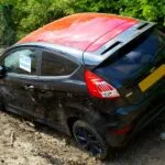 Rozbity czarny Ford Fiesta z oznaczeniem policji na wiejskiej drodze